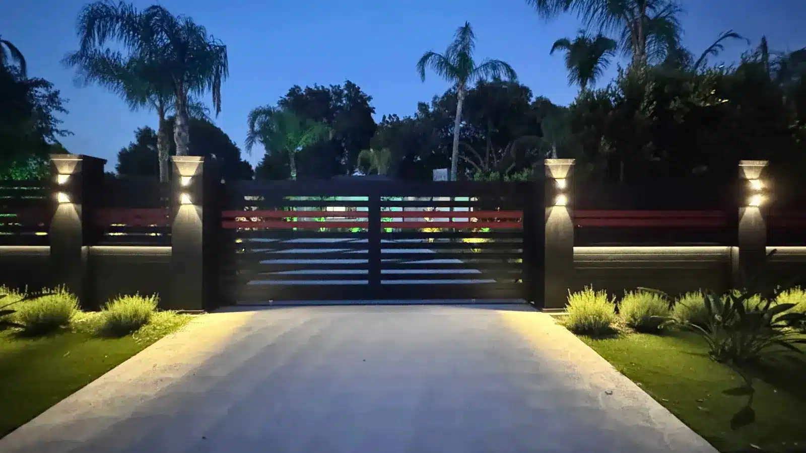 Black horizontal slat aluminum driveway gate lit at dusk between lighted pillars