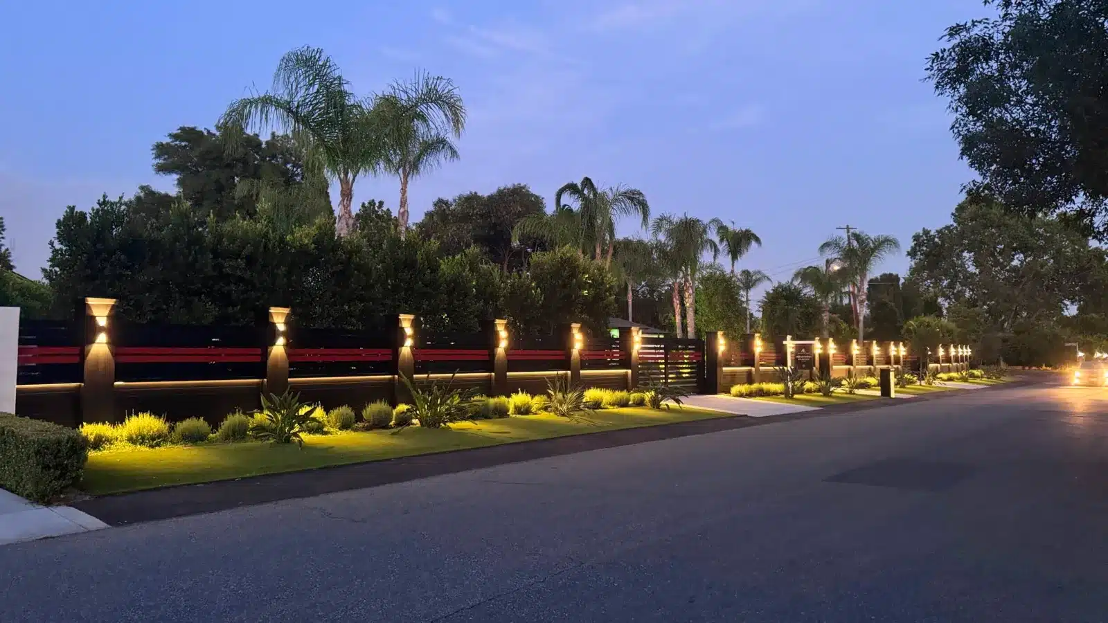 Black aluminum fence line with post lights along a hedge at dusk
